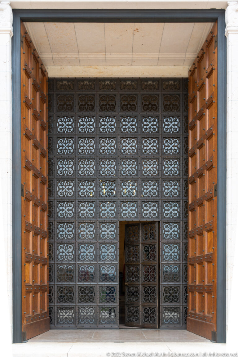 Open wooden doors frame an ornate dark metal grille with repeating floral panels and a small inset door.