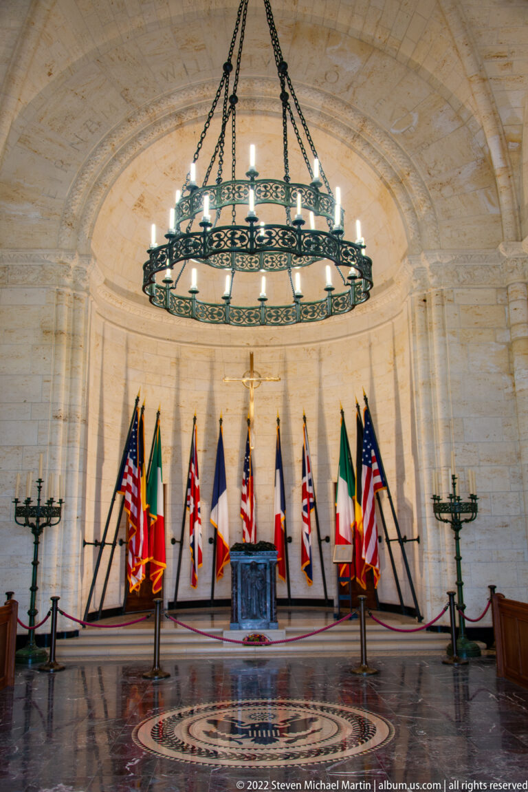 Stone chapel interior with central altar, crucifix, surrounding flags, and a large circular hanging chandelier.