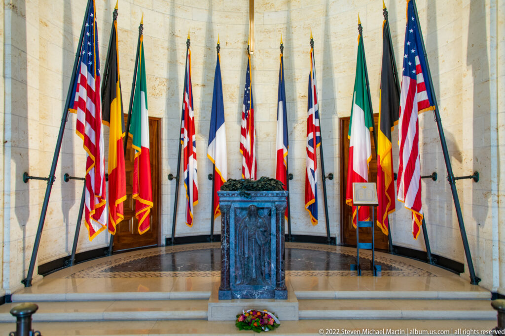 Dark stone altar on steps surrounded by American and Allied flags, with flowers placed at its base.