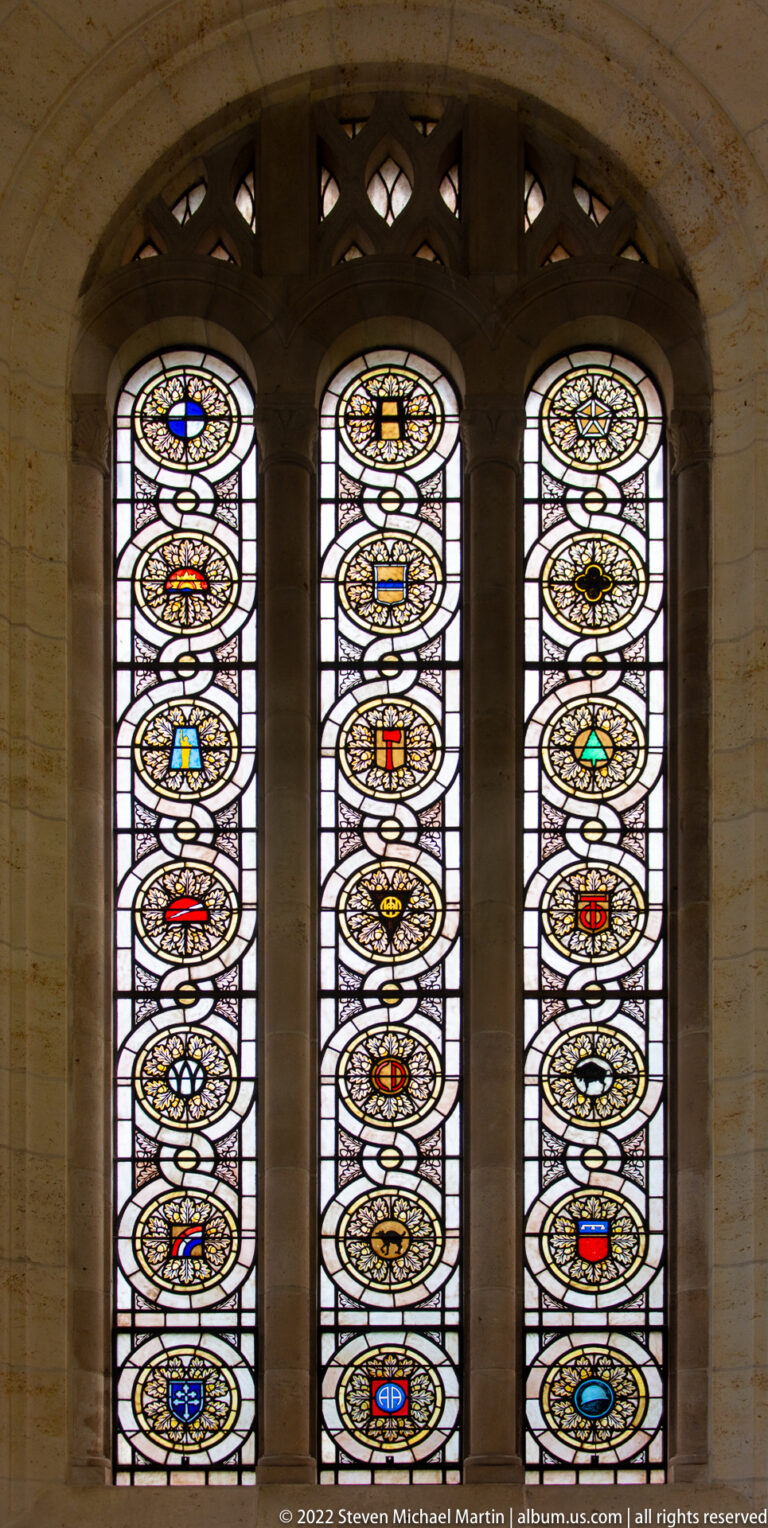 Three narrow stained-glass windows contain rows of circular military insignia and colorful heraldic emblems.