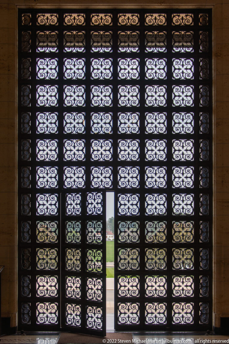Interior view of a tall patterned metal screen with daylight visible through the grille and a small open wicket door.