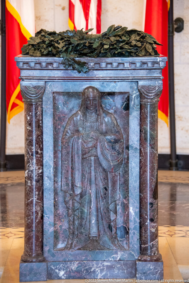 Dark stone altar with a carved robed figure on the front and a bronze laurel wreath across the top.