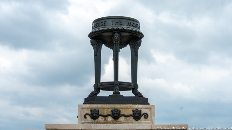 Decorated bronze circular canopy on slender columns rises above the stone memorial base under a cloudy sky.