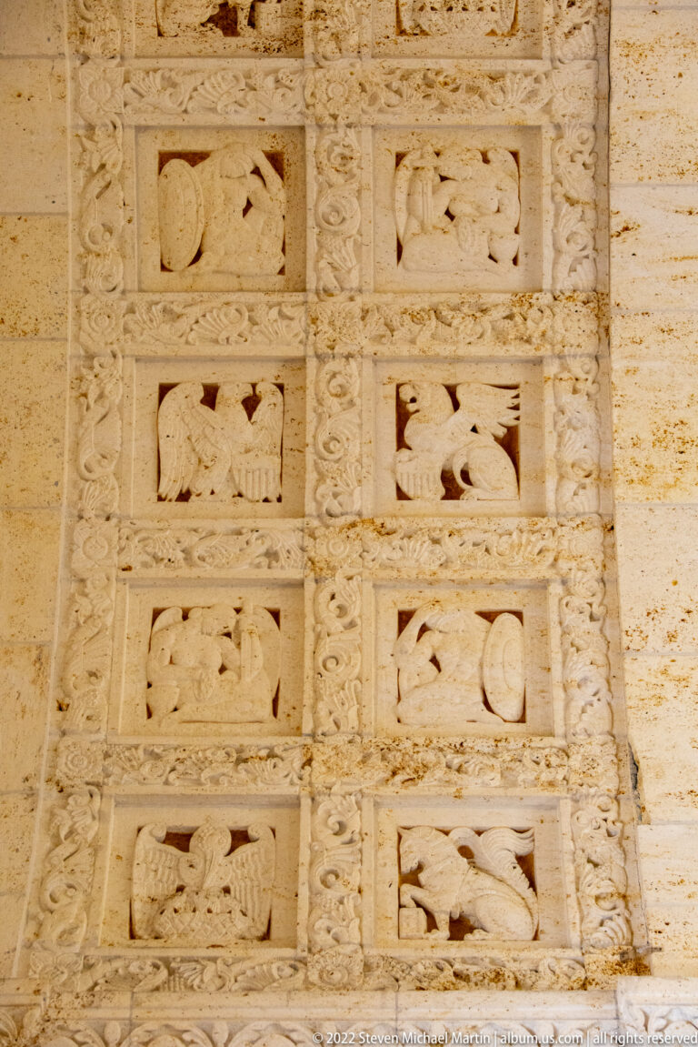 Stone wall relief with stacked rectangular panels containing birds, weapons, vessels, and other symbolic motifs.