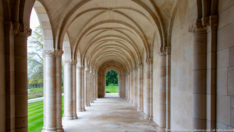Long stone arcade of repeating round arches and columns with inscribed wall panels along one side.