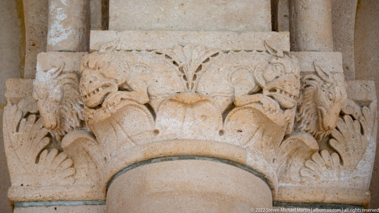 Close-up of a stone column capital carved with leaves, curled forms, and small animal or human-like heads.