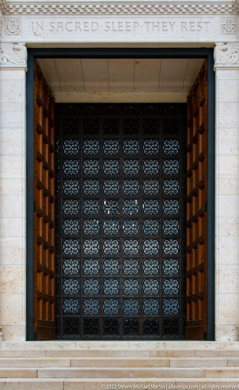 Front view of chapel entrance with open wooden doors, patterned metal screen, and carved inscription above.