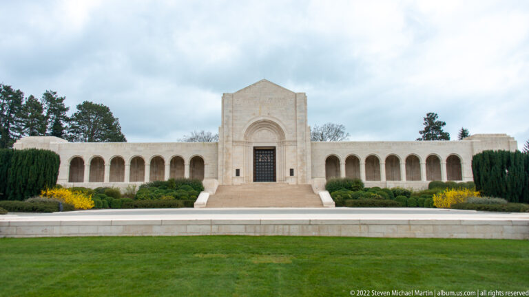 Symmetrical pale stone memorial chapel with central gabled entrance and long arcaded wings across a formal lawn.