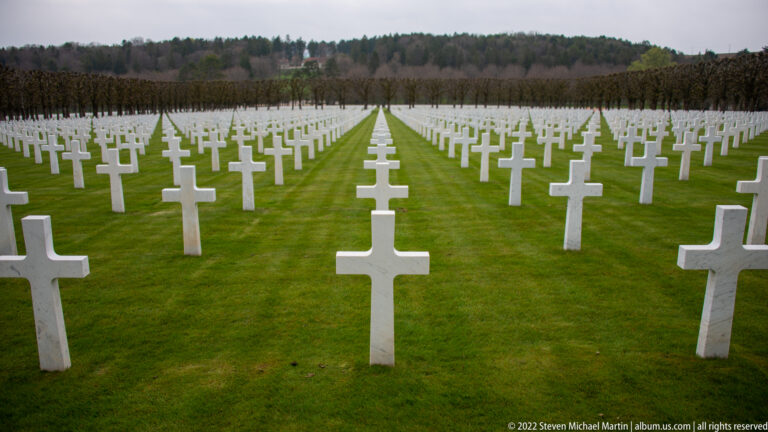 Long rows of white grave crosses recede symmetrically along a central green aisle toward trees.