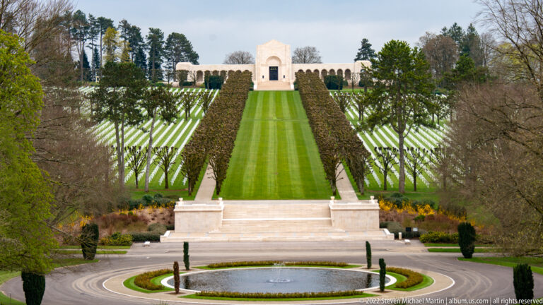 Formal cemetery axis with fountain, terraces, green lawns, rows of white graves, and chapel on the hill.