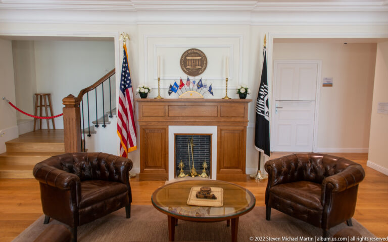 Visitor room with wooden fireplace, two leather chairs, round table, American flag, and black commemorative flag.