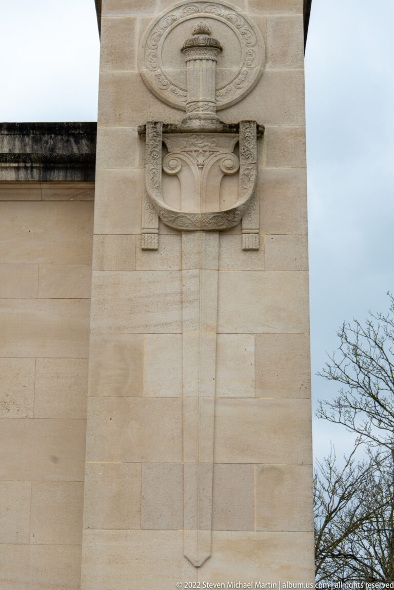Close-up of pale stone relief with a tall central urn-like motif, scrolls, and circular ornament.