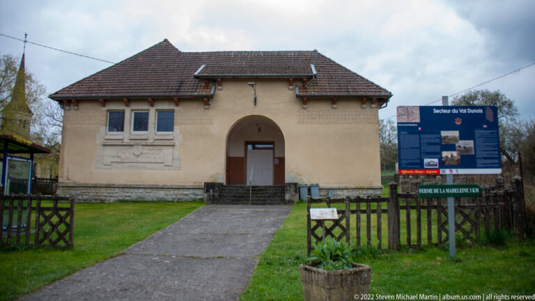 Small pale building with tiled roof and arched doorway beside a large outdoor interpretive sign.
