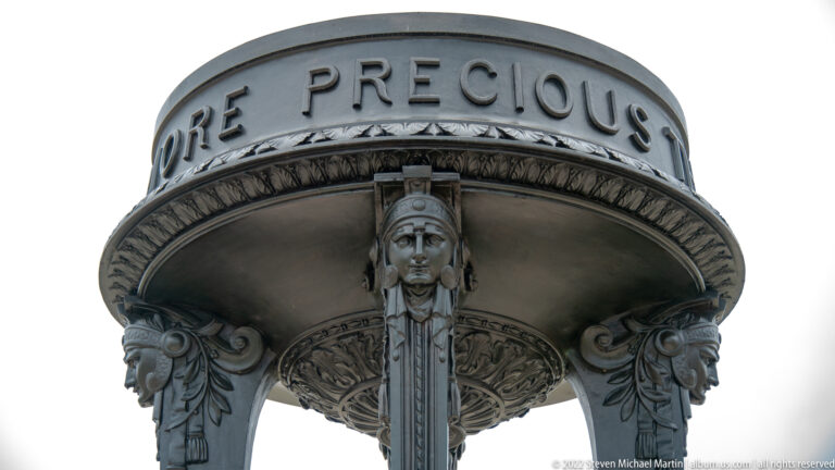 Close-up beneath a bronze memorial canopy showing raised lettering, floral ornament, and a sculpted female support.