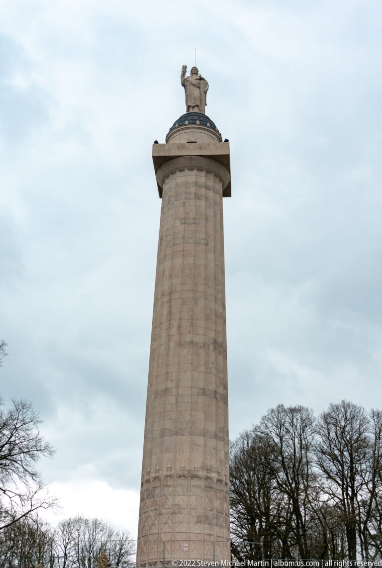 Tall tapering stone monument with carved lower panels and a sculpted top, seen from below against cloudy sky.