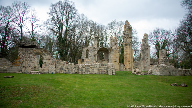 Ruined stone walls, arches, and tall broken piers stand on grass beneath leafless trees.