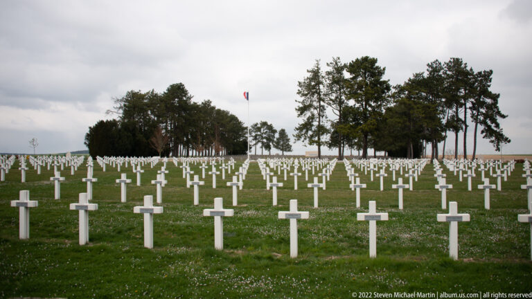 Rows of white grave crosses cover a broad lawn with trees and a small French flag in the distance.