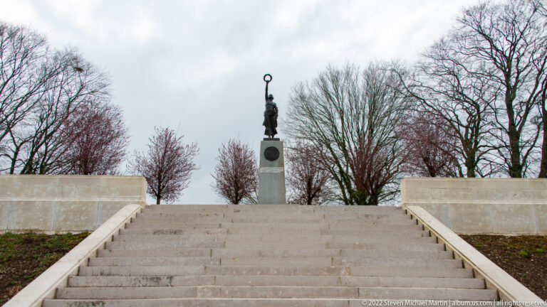 Wide stone steps lead upward between low walls to a standing figure on a tall pedestal.