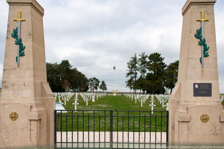 Stone cemetery gate piers with sword-and-laurel decoration frame rows of white crosses beyond a black gate.