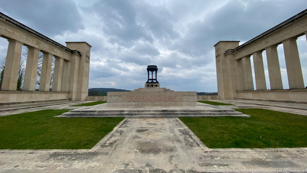 Central stone monument on a paved terrace between matching open colonnades beneath a cloudy sky.