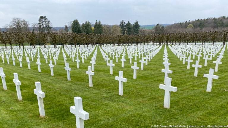 Wide elevated view over hundreds of white grave crosses arranged in long rows across green lawns.