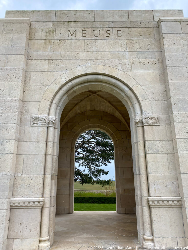 Large pale stone arch inscribed Meuse frames a green lawn and tree beyond.