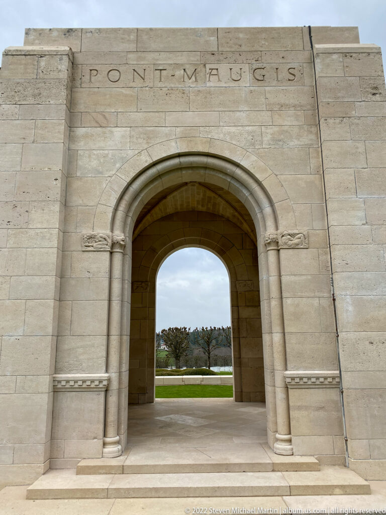 Large pale stone arch with engaged columns frames lawn and trees beneath a partial carved place-name inscription.