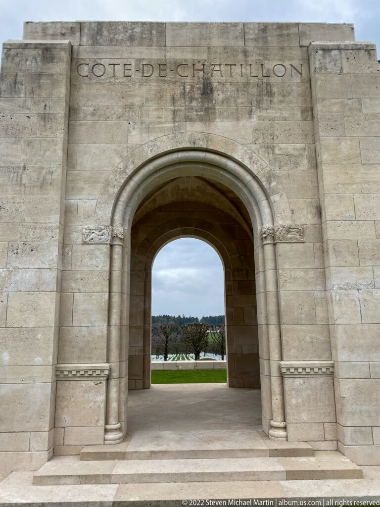 Large round stone arch with partial carved place-name above, framing clipped trees and lawn beyond.