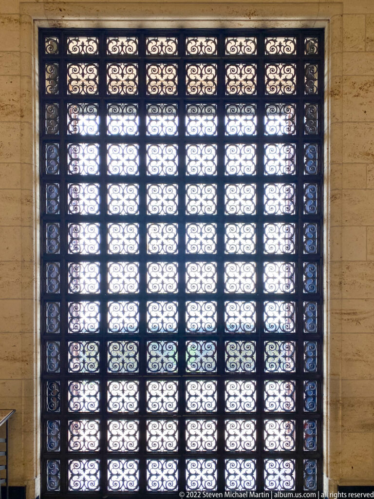 Backlit dark metal grille fills a tall opening with rows of repeating floral and scroll patterns.