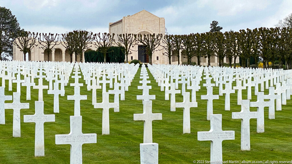 Rows of white grave crosses recede along a central axis toward the pale memorial chapel behind pollarded trees.