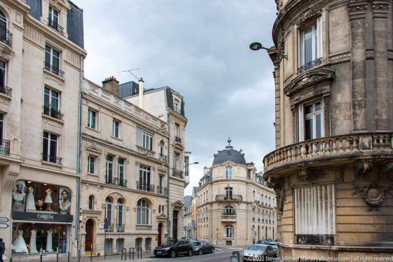 Central Reims street corner with pale stone buildings, balconies, shopfronts, parked cars, and a rounded corner façade.