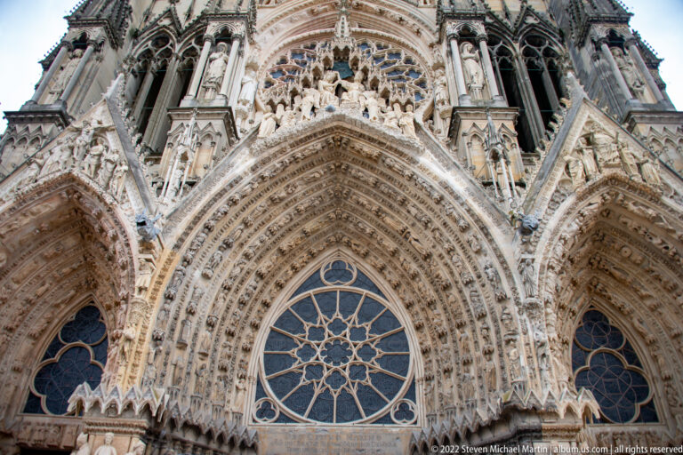 Upward view of Reims Cathedral’s west façade with rose window, pointed portals, tracery, sculptures, and tower bases.