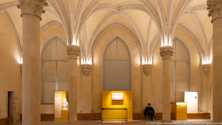 Gothic-vaulted museum gallery with pointed wall recesses, display cases, and one seated visitor.