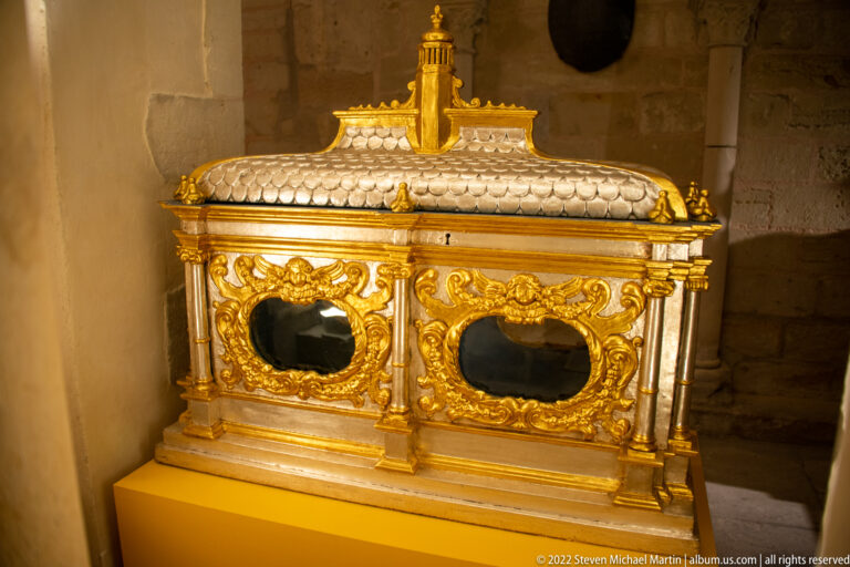 Rectangular gilt reliquary casket with two dark glazed openings, miniature roof, pilasters, and finial.
