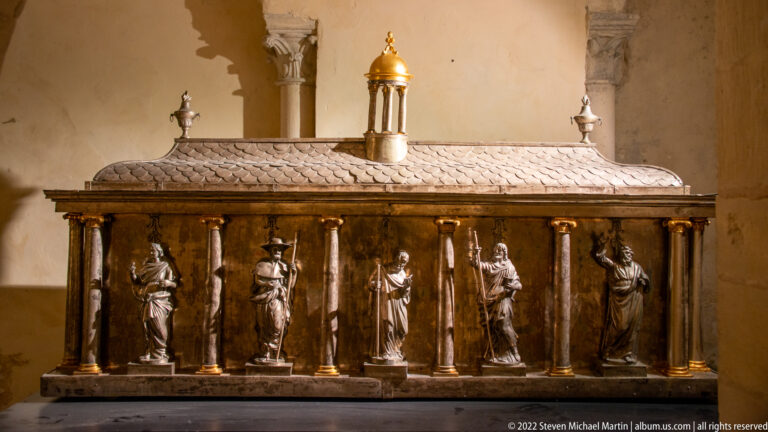 Long metal reliquary shrine with standing figures in columned bays beneath a patterned sloping roof.