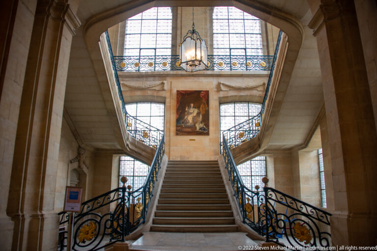 Grand stone staircase with ornate blue-and-gold railings, tall windows, hanging lantern, and central painting.