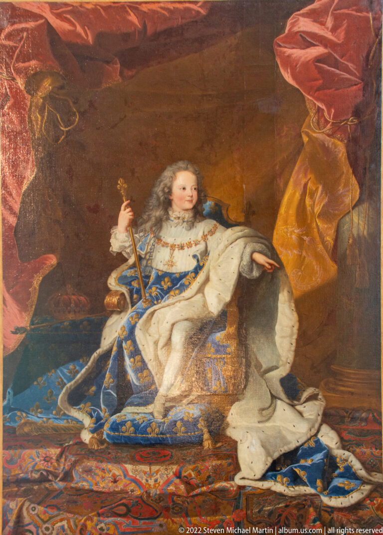 Young prince in blue fleur-de-lis and ermine robes holding a ceremonial staff before red drapery.