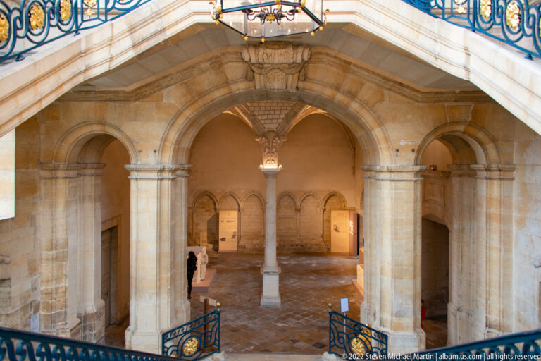Stone-vaulted hall beneath a grand staircase, with rounded arches, central column, and blue-and-gold railings above.