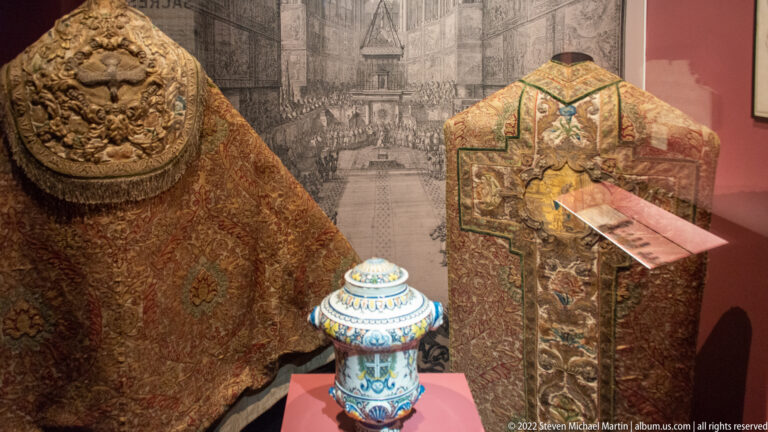 Two richly embroidered church vestments displayed beside a decorated lidded ceramic vessel.