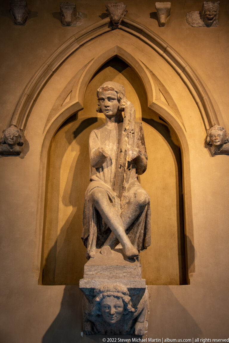 Weathered seated stone figure beneath a pointed niche, with one leg crossed and a carved head below.