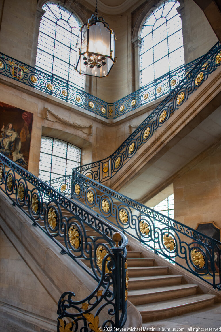 Diagonal stone staircase with ornate blue wrought-iron railings, gilded medallions, tall windows, and hanging lantern.
