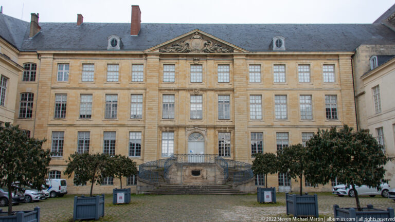 Symmetrical former abbey façade with central pediment, broad curving stair, rows of windows, and courtyard trees.