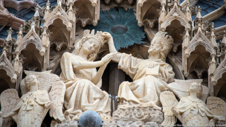 Close view of two carved Gothic figures gesturing toward each other beneath canopies on Reims Cathedral’s façade.
