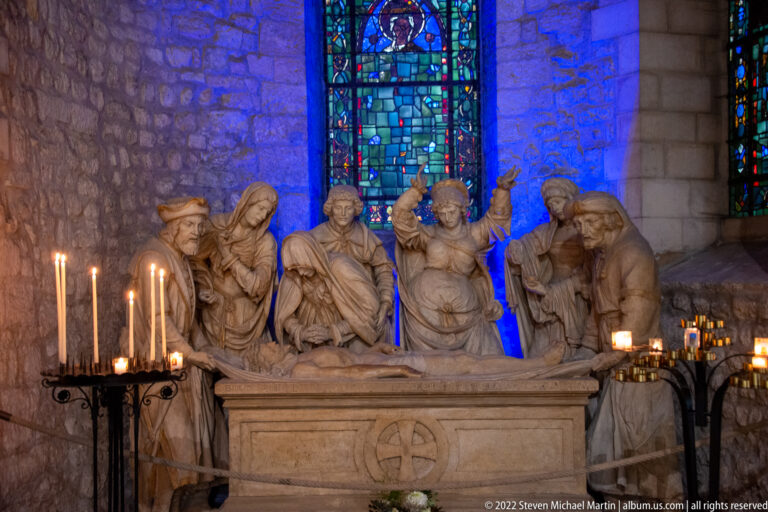 Stone Lamentation group with mourners surrounding the dead Christ beside candles and blue stained glass.