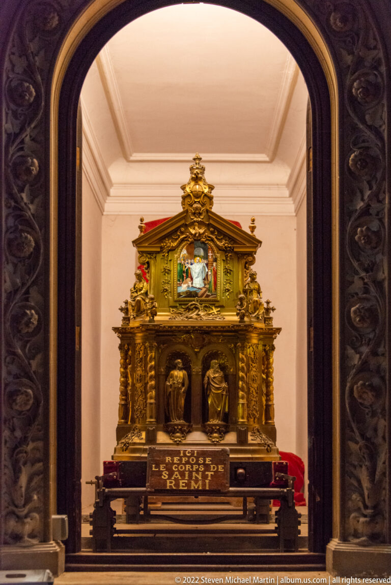 Elaborate gilded reliquary with miniature architecture, reliefs, and a small colored scene in a chapel niche.