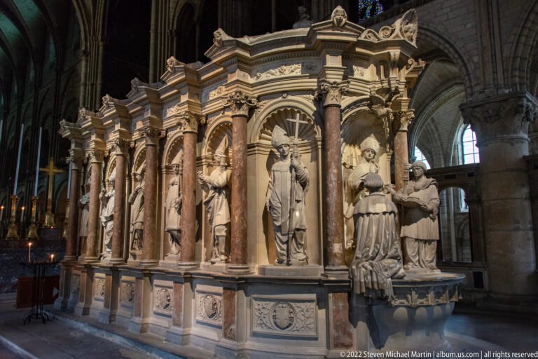 Oblique view of a monumental shrine with saint statues, marble columns, and a kneeling figure at one end.