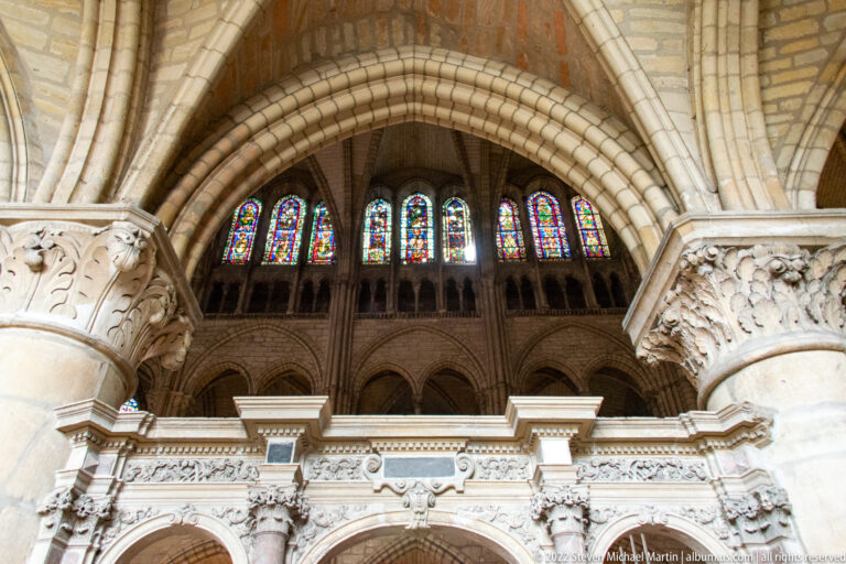 Upward interior view of heavy stone arches, carved capitals, pointed openings, and stained-glass windows.