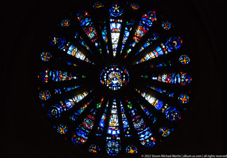 Dark-framed rose window with multicolored radial stained-glass panels and a small central medallion.