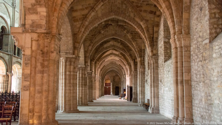 Long basilica aisle with repeating stone arches, heavy piers, ribbed vaults, and a distant doorway.