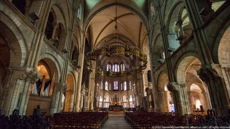 Wide view down the Basilica of Saint-Remi nave toward the choir, with chairs, stained glass, and large chandelier.
